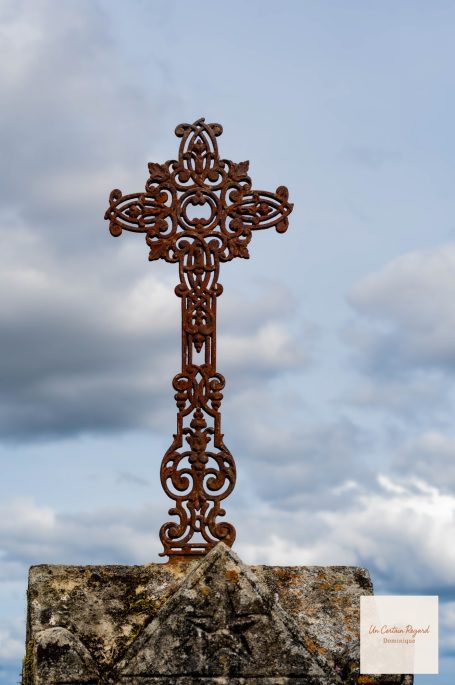 Croix en fer forgé sur un socle, contre un ciel nuageux.